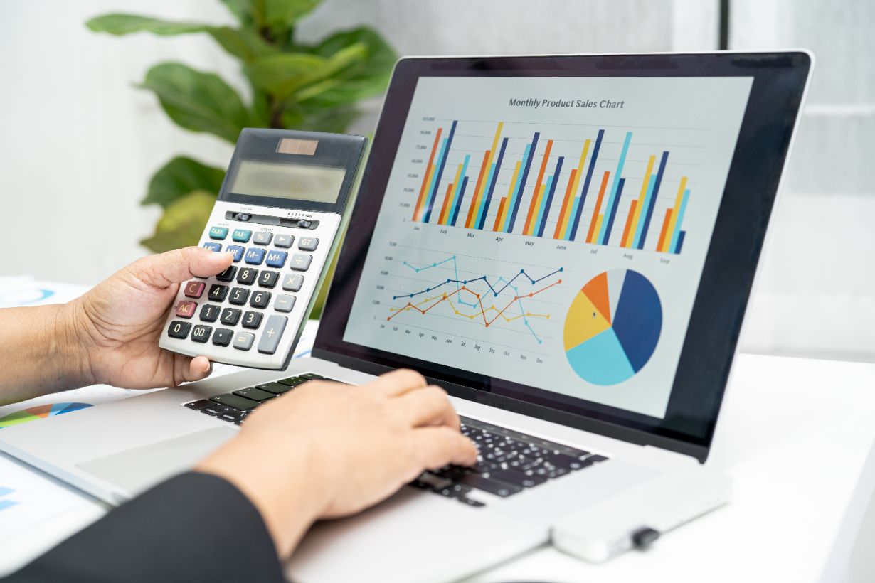 A person is using a calculator alongside a laptop showing a detailed monthly product sales chart, complete with bar, line, and pie graphs, while ensuring compliance with ATO Guidelines 2024 for accurate record-keeping.