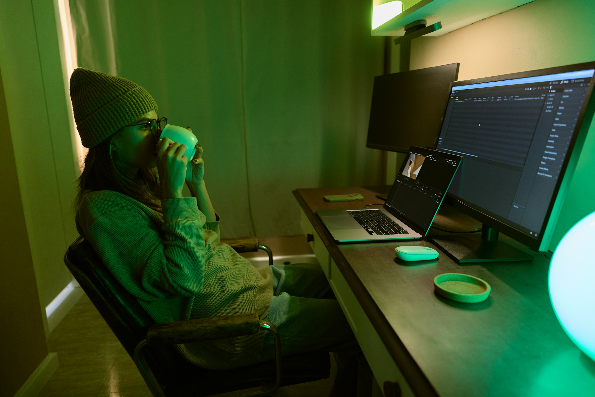 Amidst green lighting, a freelancer in a beanie and glasses sips a drink while working on a dual-monitor setup. Immersed in tasks, thoughts of balancing creativity with responsibility like tax duties occasionally surface.