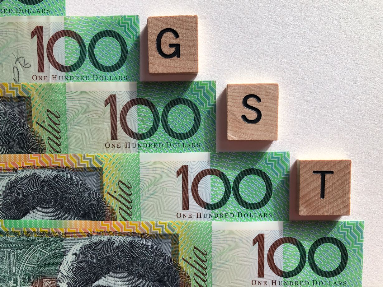 Australian 100 dollar notes are arranged in a staircase pattern, with wooden tiles labeled "G," "S," and "T" on top, symbolizing the essential guide for Australian businesses in navigating GST.