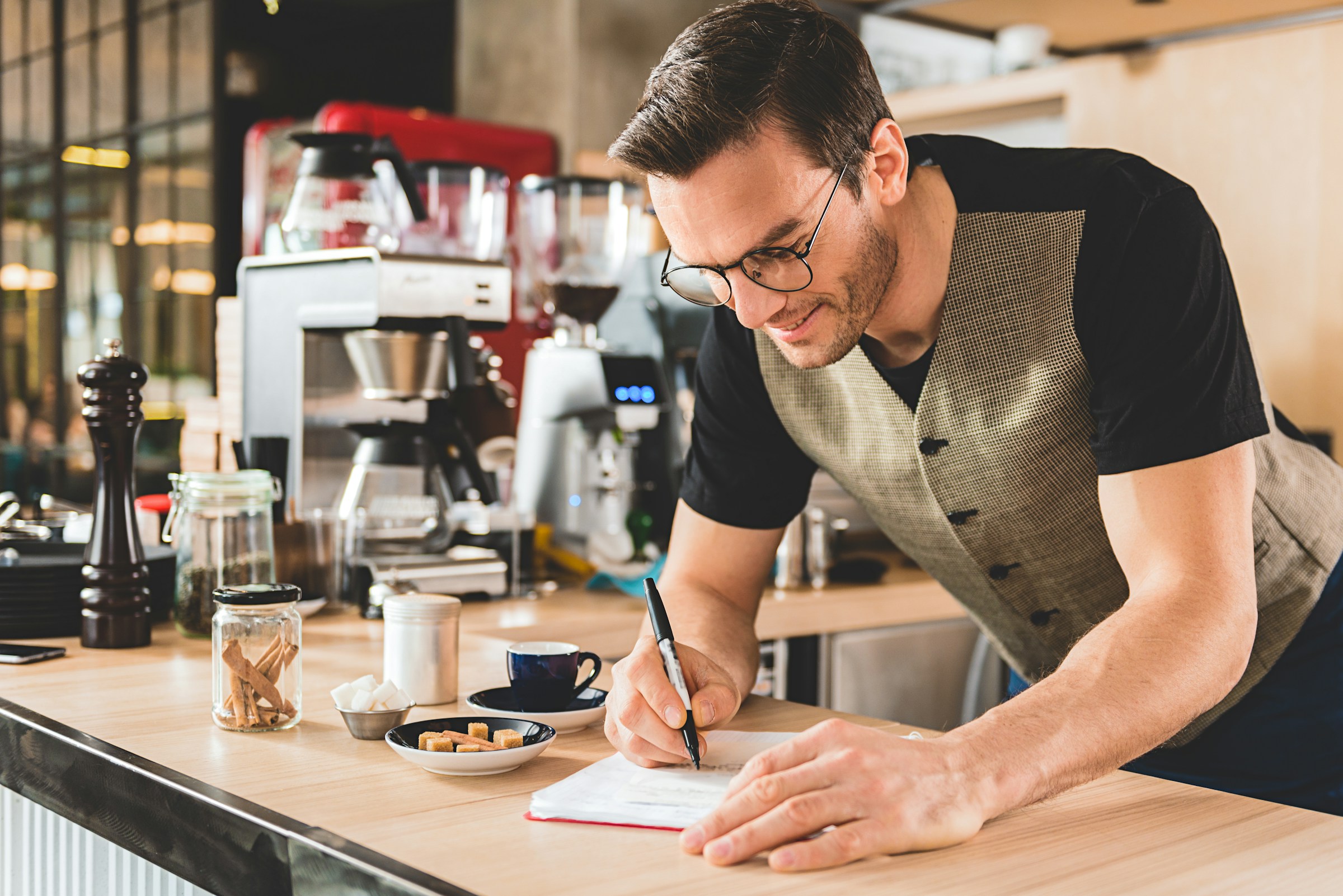 In a cozy coffee shop, a man pens down notes on "How to Pay Yourself as a Business Owner," surrounded by the comforting aroma of freshly brewed coffee and an array of brewing tools.