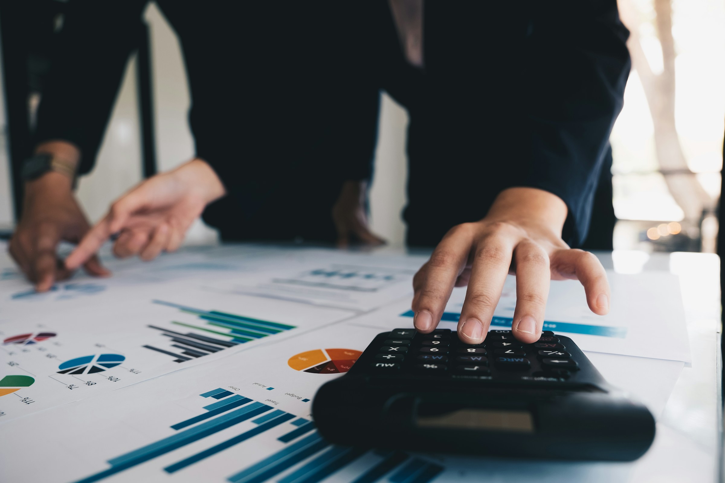 Two people in business attire work on financial graphs and charts on a table, ensuring payroll compliance. One uses a calculator, while the other points at a chart.