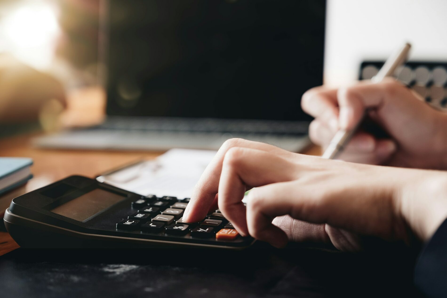 A person diligently ensures payroll compliance, using a calculator and writing with a pen on paper, with a laptop in the background.