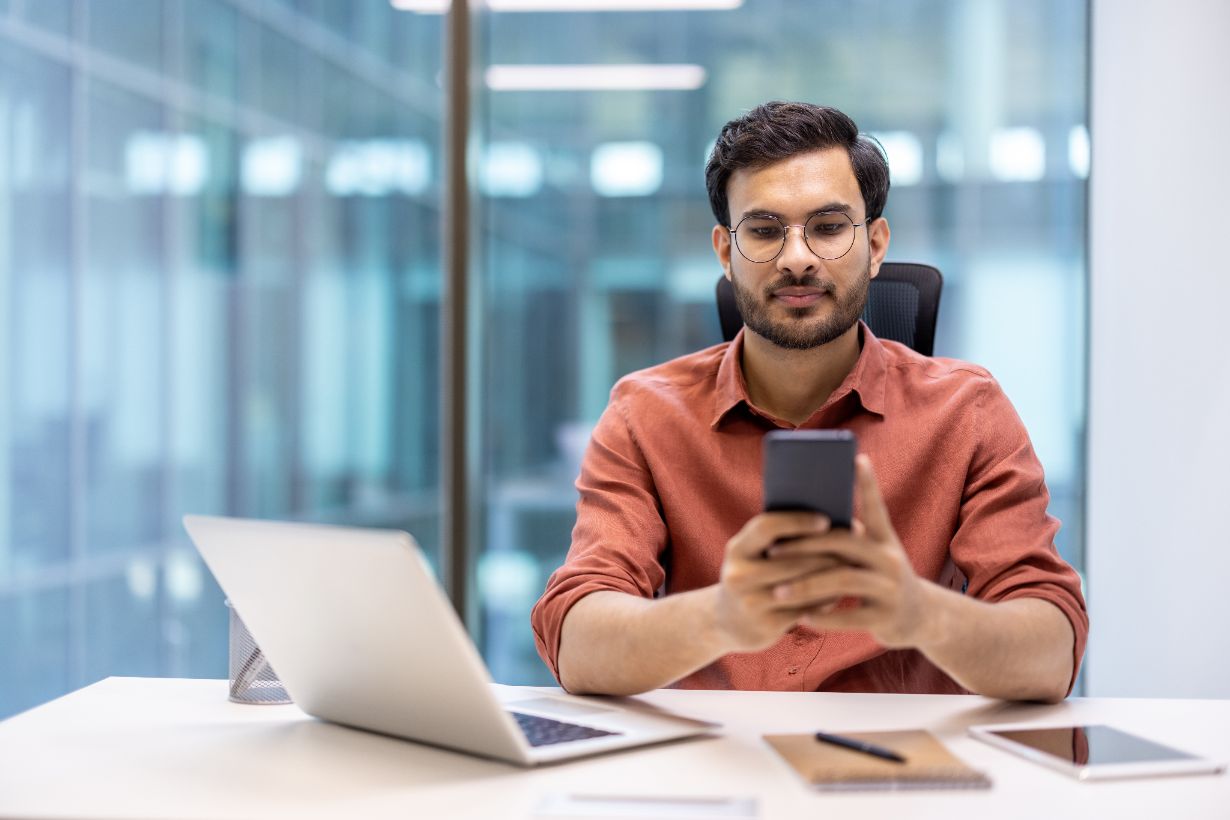 In an office setting, a man wearing glasses and a brown shirt sits at his desk using a smartphone. Surrounded by a laptop, notebook, and pen, he appears ready to guide Australian businesses on GST matters.