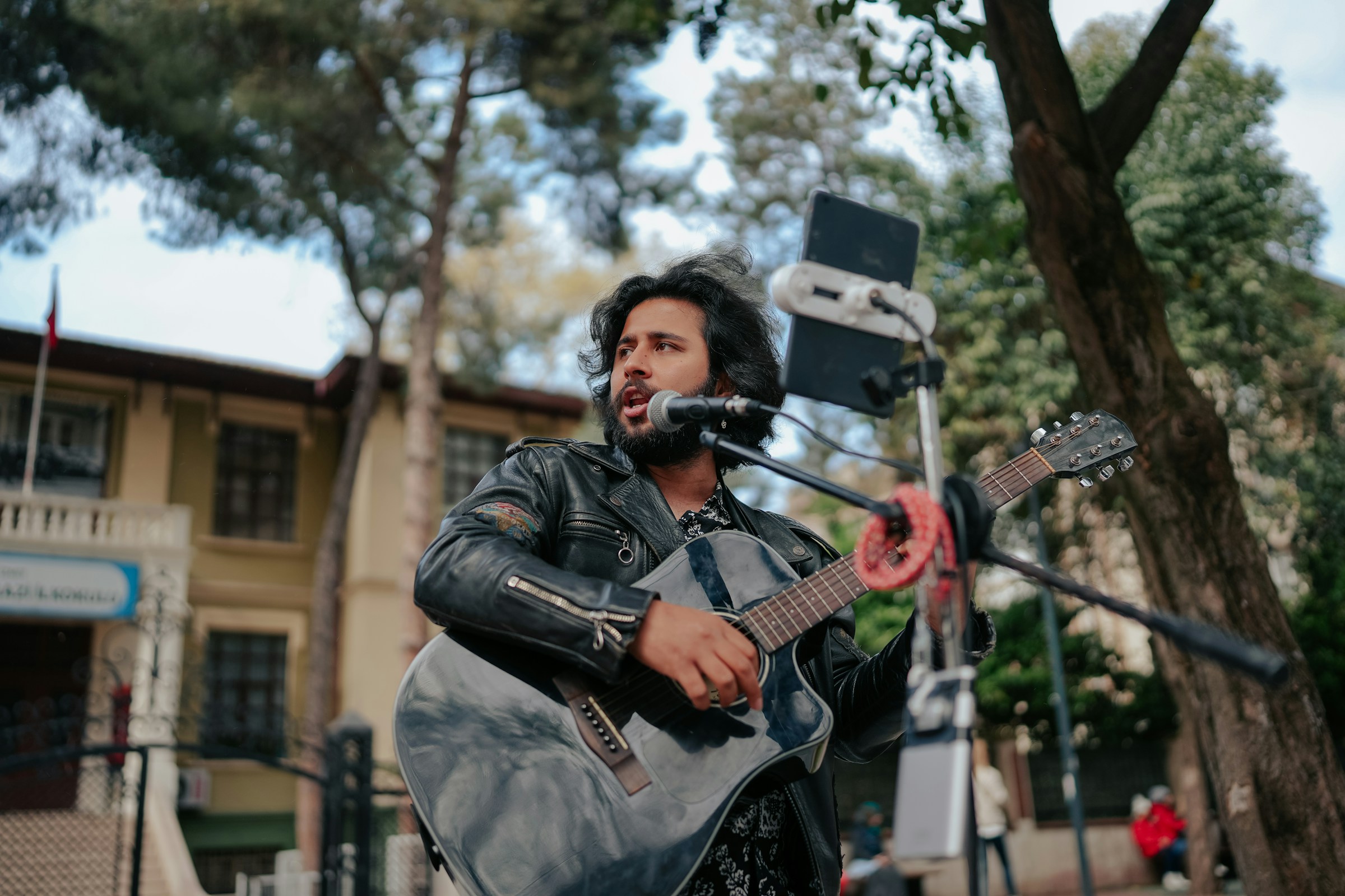 A musician skillfully plays an acoustic guitar and sings outdoors using a microphone, surrounded by trees and a building in the background. If you're curious about what musicians can claim on tax, this setting surely hints at potential deductions for equipment and performance spaces.