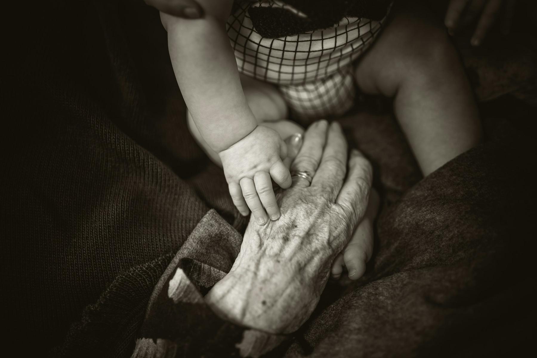 A baby's hand touches an elderly person's hand, showcasing generational connection. The elderly hand is resting on a blanket.