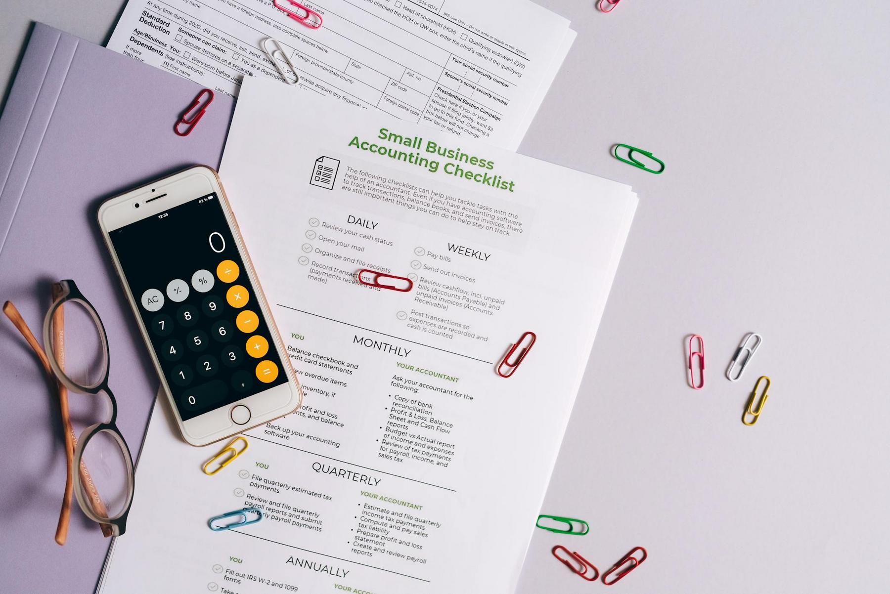 Small business accounting checklist on a desk with a smartphone calculator app, eyeglasses, paperwork, a folder, and scattered colorful paper clips.