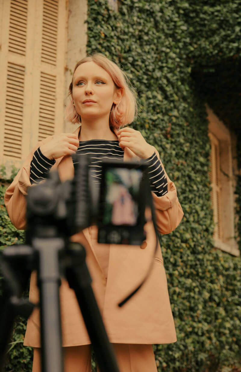 A person with pink hair adjusts their striped shirt while standing in front of an ivy-covered wall. The scene is being recorded by a camera on a tripod in the foreground.