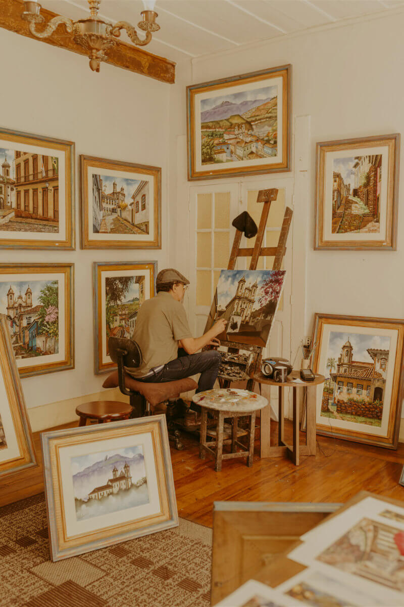 An artist paints at an easel in a room filled with framed landscape and building paintings. He is seated on a stool, surrounded by various art supplies and completed works displayed on the walls.