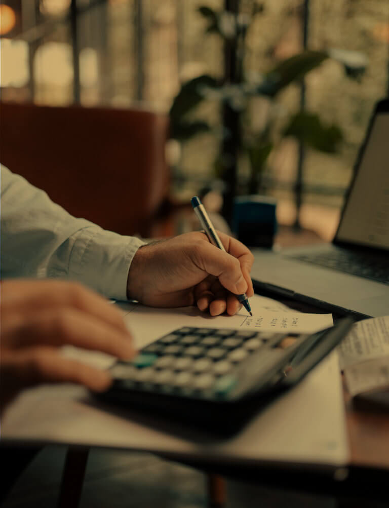 A person's hands are shown using a calculator and writing on a piece of paper, with a laptop and papers on the table in the background.