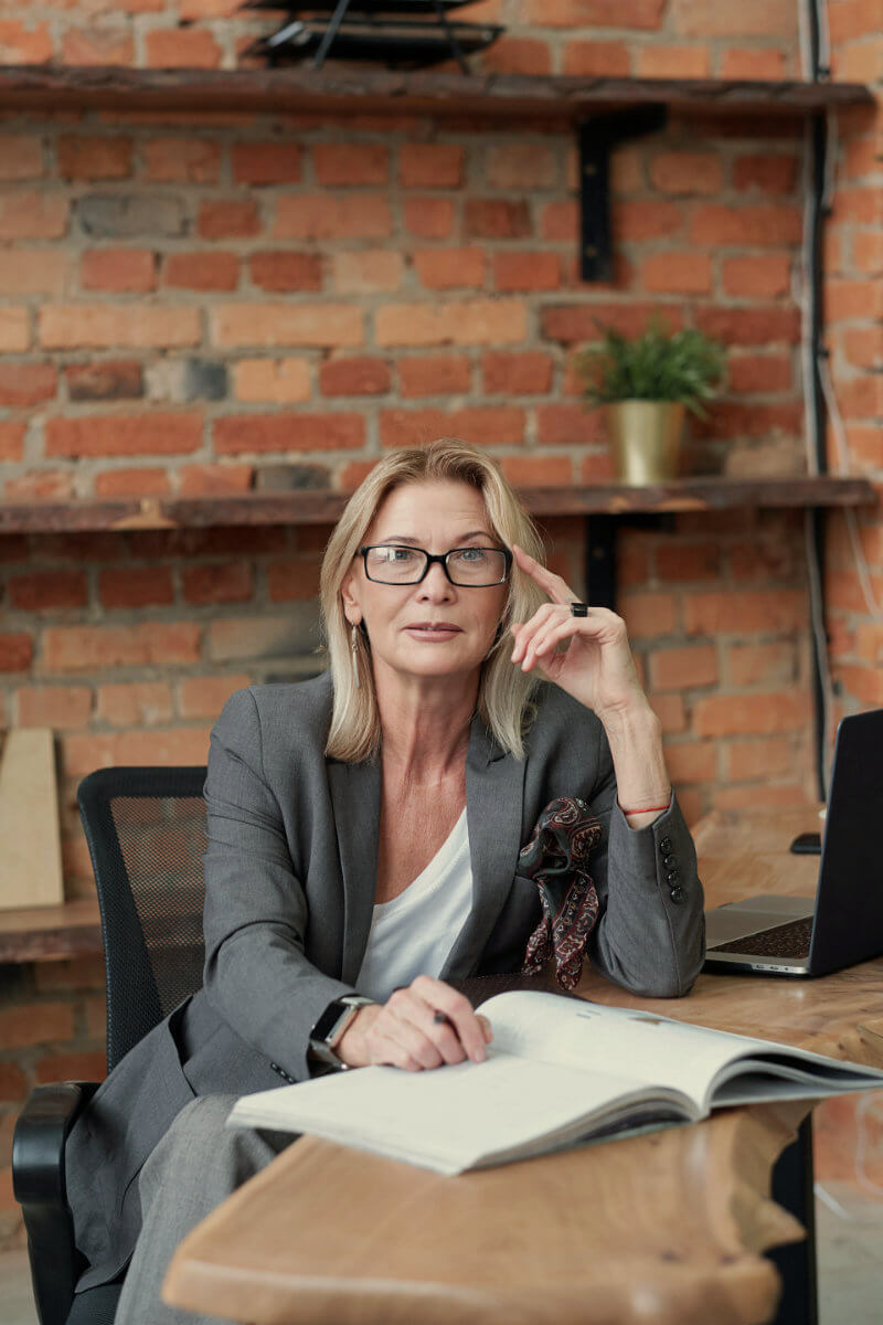 A woman with glasses, wearing a grey suit, is seated at a desk in front of a brick wall, looking directly at the camera with an open book in front of her and a laptop on the side.