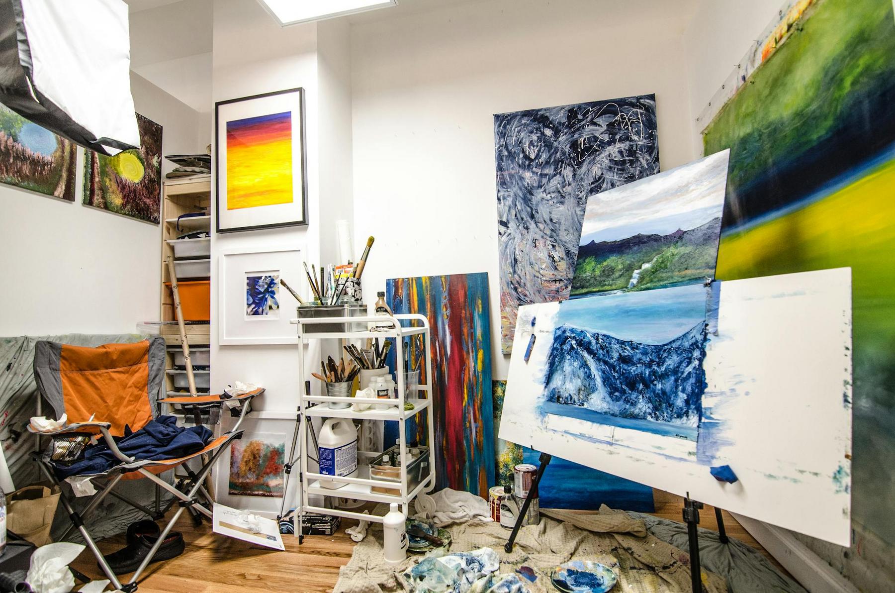 An artist’s studio with canvases, paintings, brushes, and art supplies; a painting of waves sits on an easel, surrounded by various artworks and a folding chair.