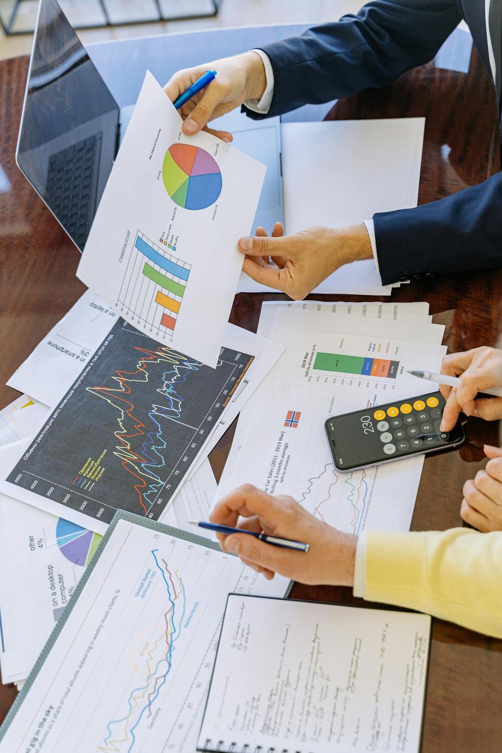 Two people analyzing printed charts and graphs on a desk, with a calculator, pen, notebook, and laptop visible.