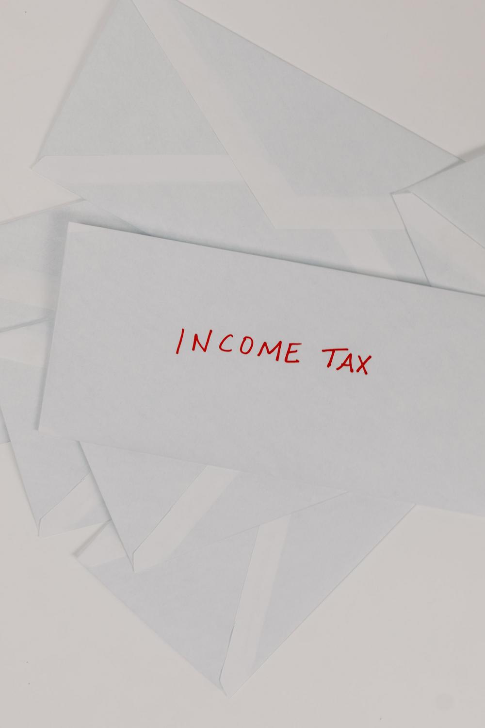 A white envelope labeled "INCOME TAX" in red marker lies on top of several other blank white envelopes on a light surface.