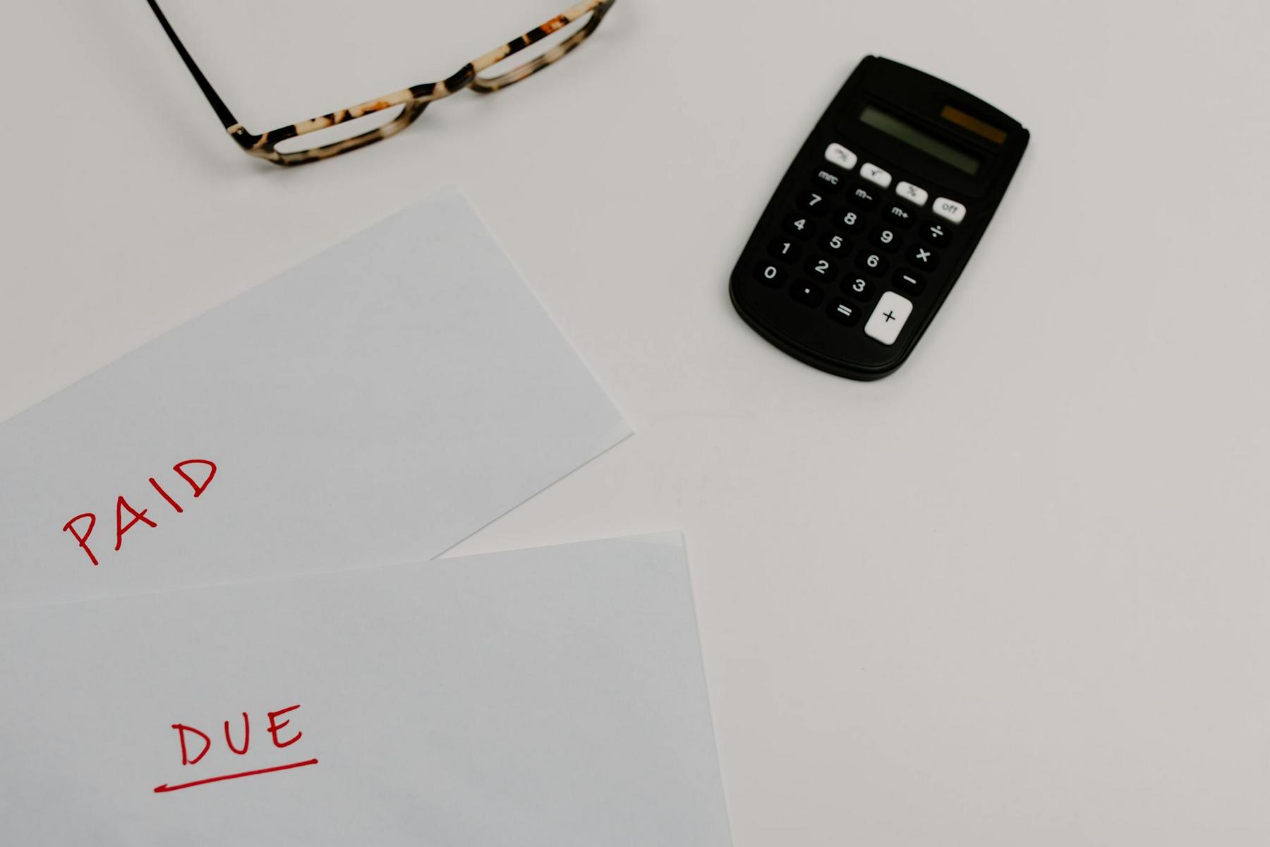 Two envelopes labeled "PAID" and "DUE" in red ink are on a white surface next to a pair of eyeglasses and a black calculator.