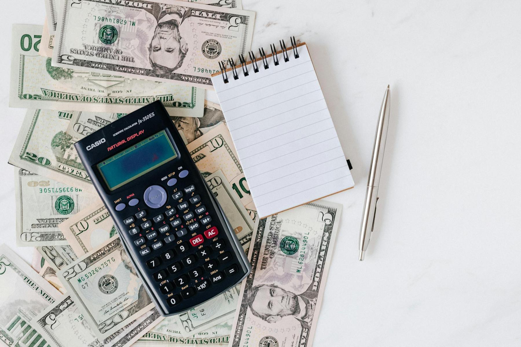 A scientific calculator, a notepad, and a pen are placed on top of scattered US dollar bills on a white surface.
