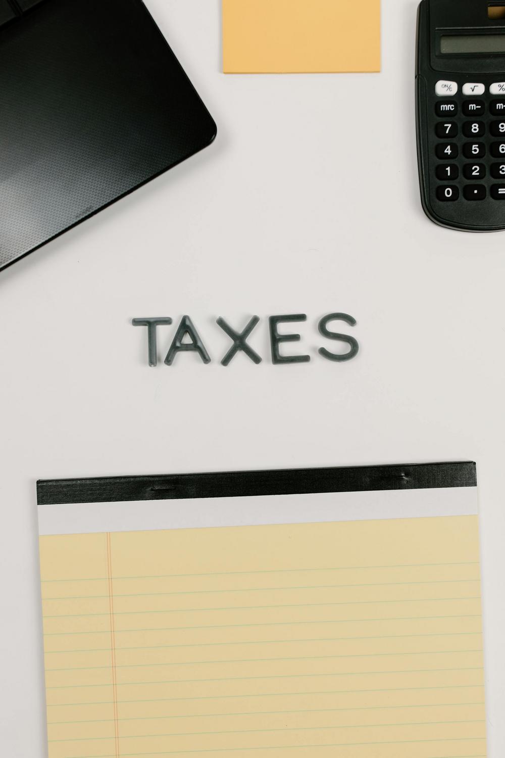A notepad, a calculator, a laptop, and a sticky note surround the word "TAXES" spelled out in letters on a white surface.
