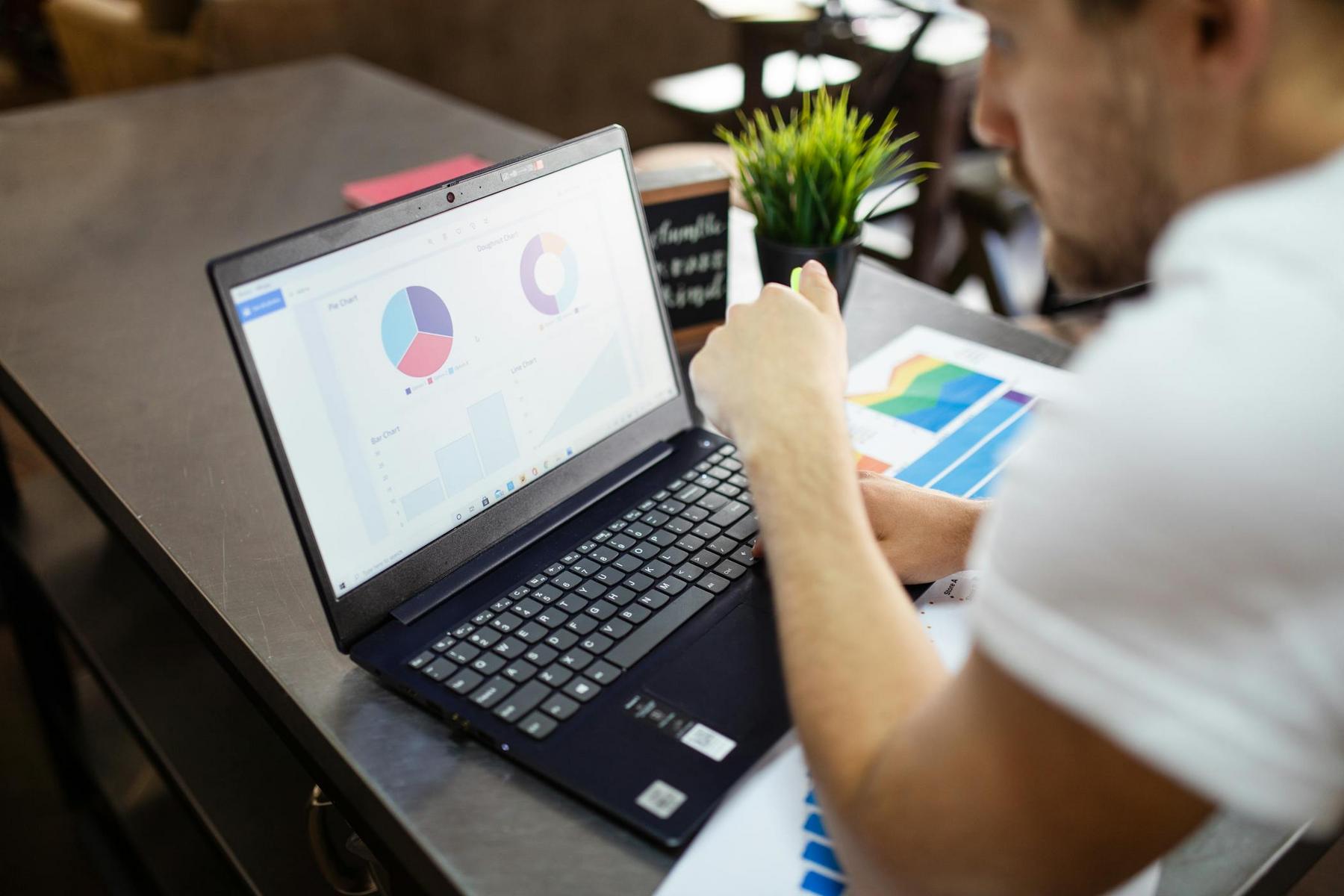 A person viewing charts and graphs on a laptop screen while working at a desk with documents and a small plant.