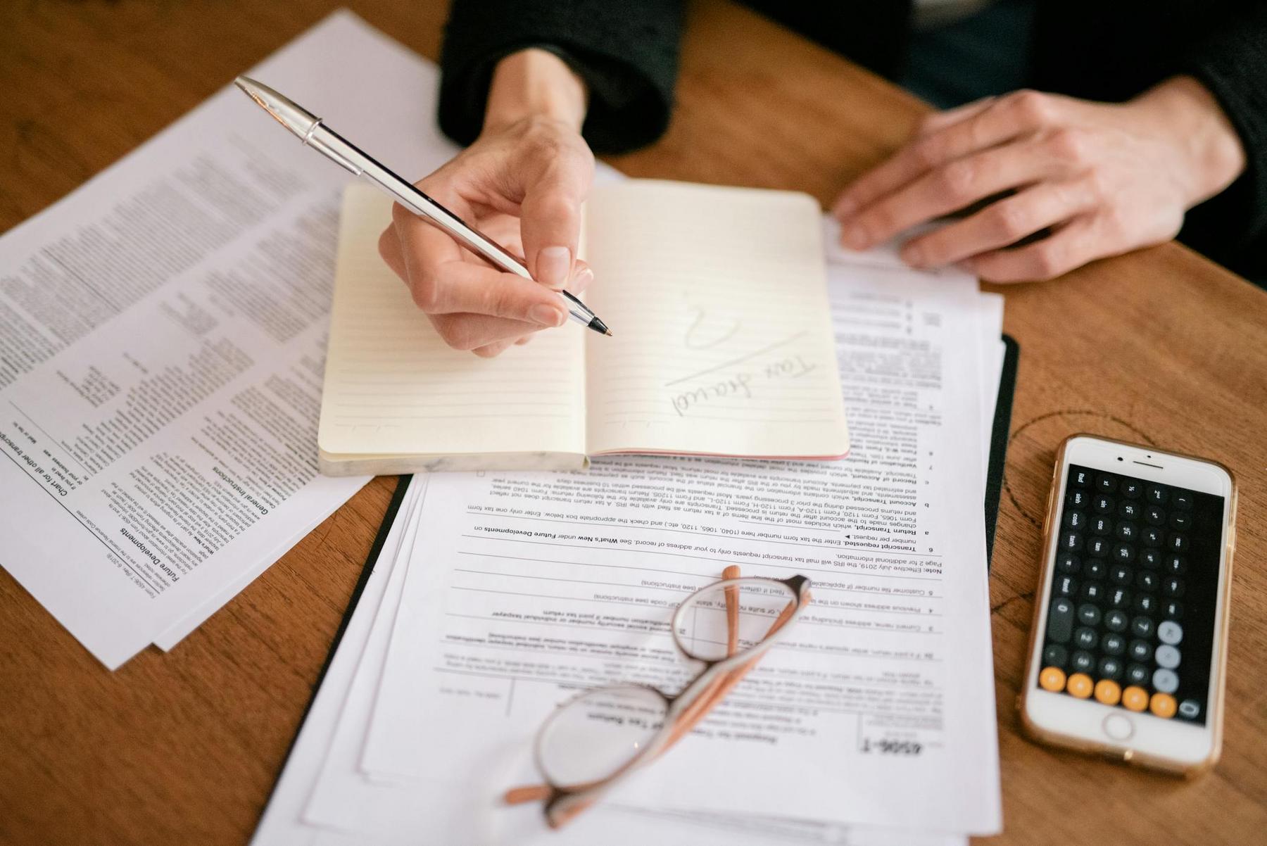 A person writing in a notebook surrounded by paperwork, a pair of glasses, and a smartphone with the calculator app open on a wooden table.