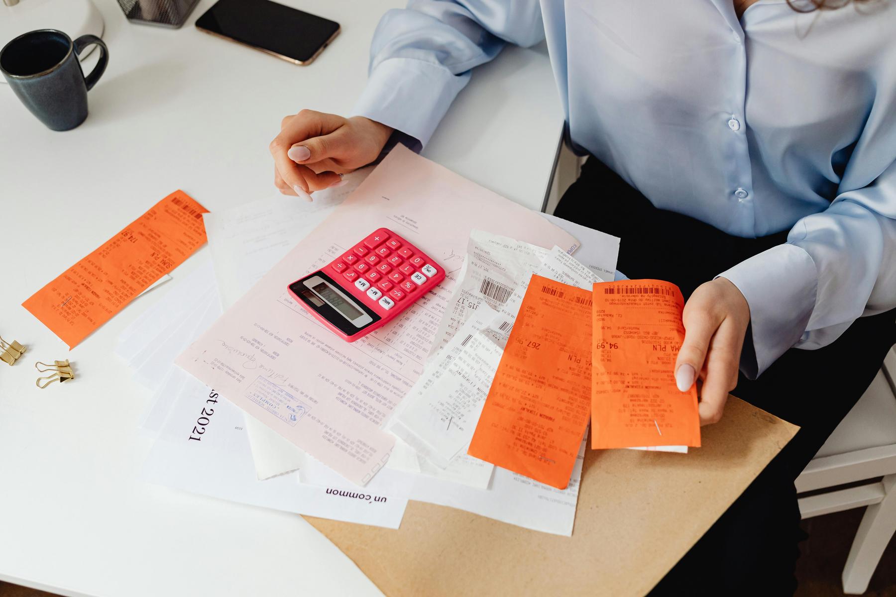 A person sits at a desk with a pink calculator, receipts, documents, and a cup of coffee, appearing to review or organize financial paperwork.