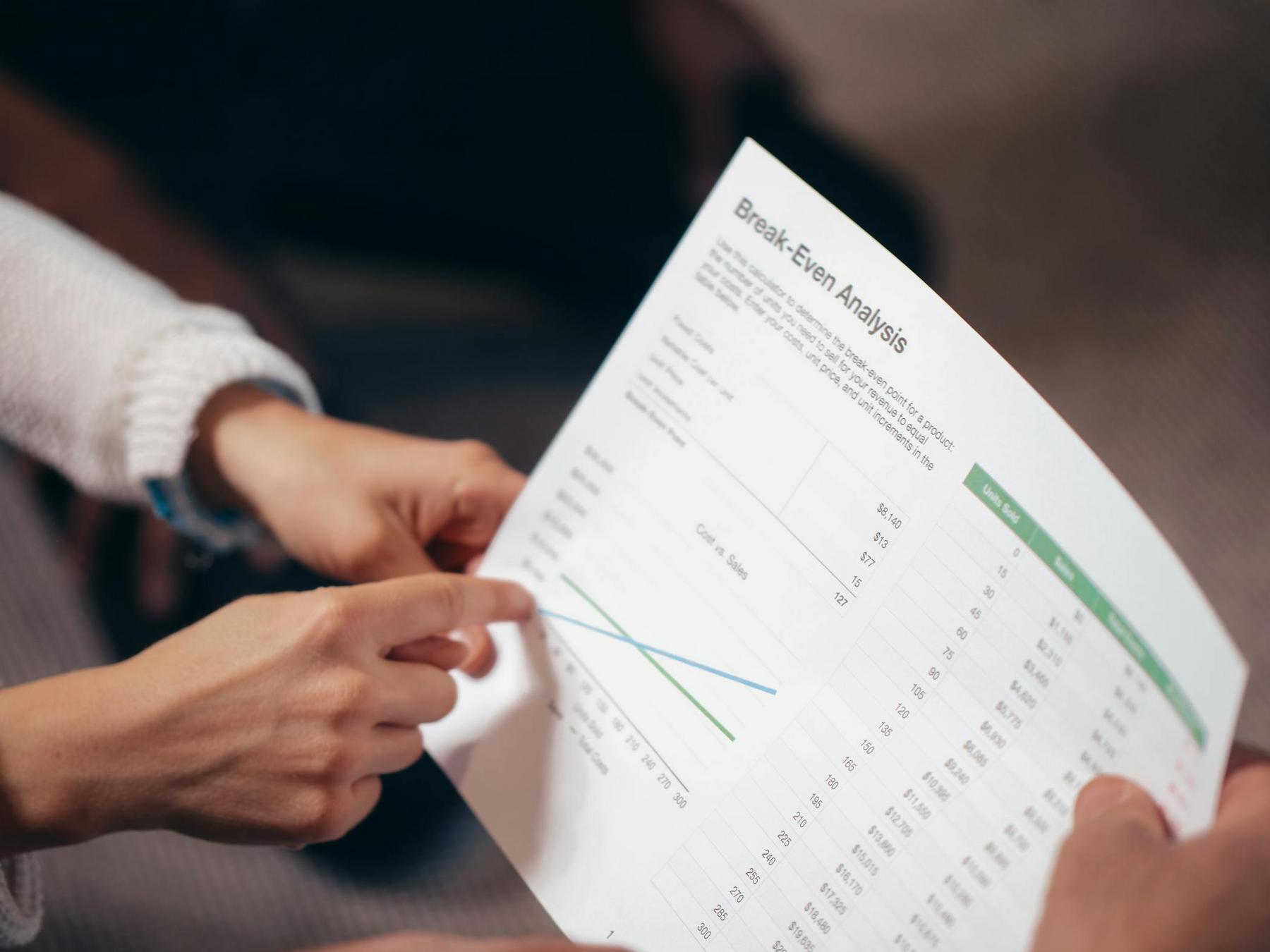Two people review a printed break-even analysis chart with data, graphs, and tables; one person points at the graph on the paper.
