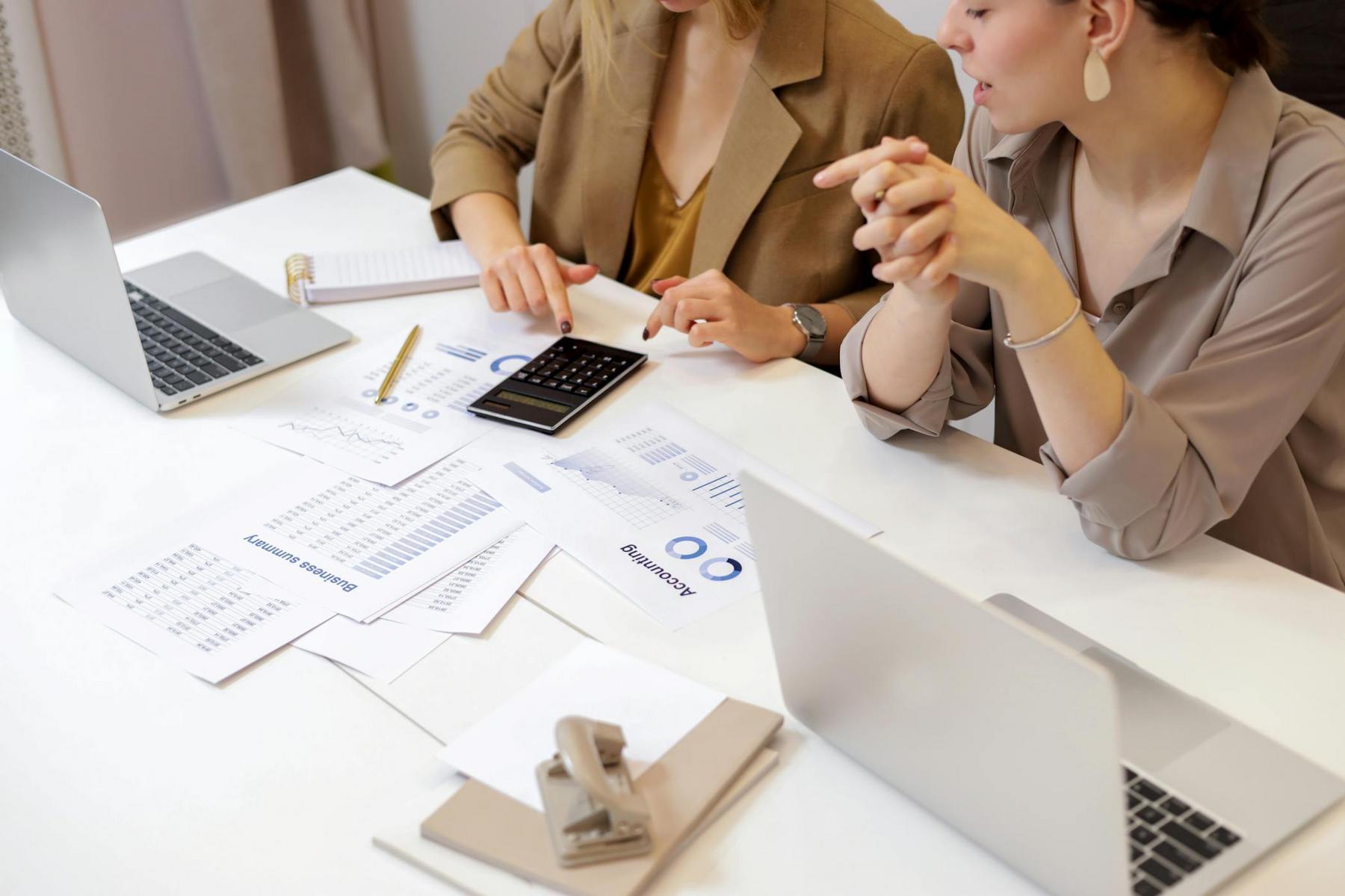 Two people sit at a desk with laptops, documents, a notepad, a calculator, and a pen, appearing to discuss business charts and financial data.