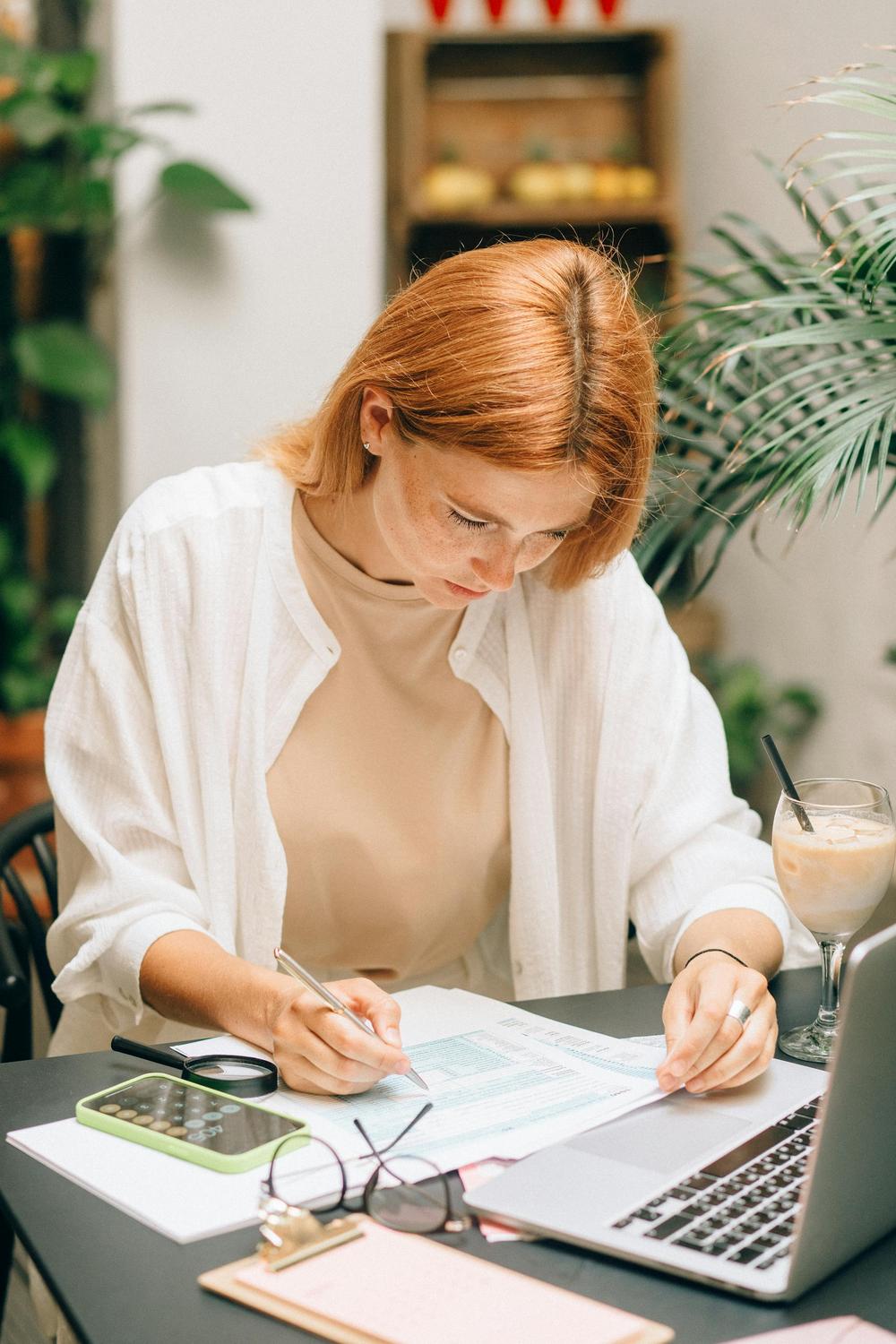 A person with red hair sits at a desk, filling out paperwork with a pen next to a laptop, smartphone, calculator, and iced drink in a bright room with plants.