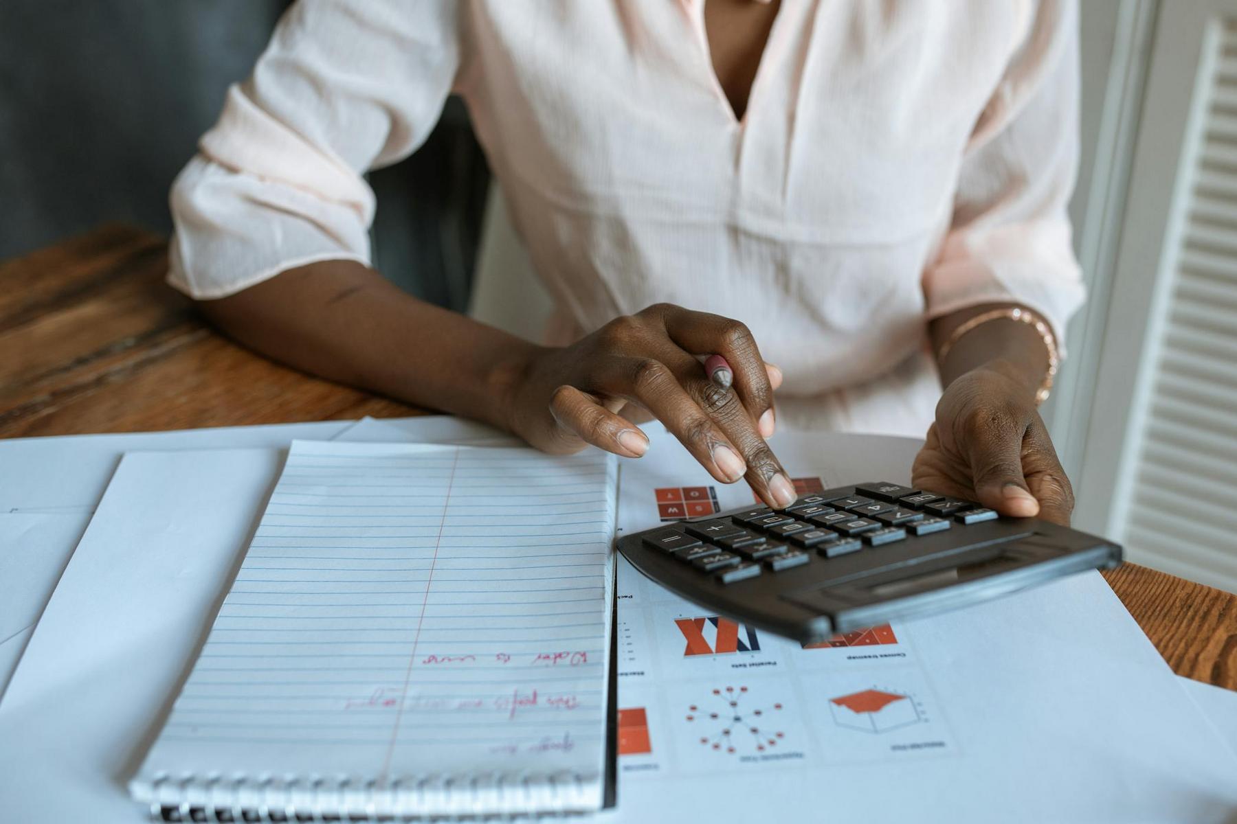 A person uses a calculator at a desk with a notepad and printed charts, appearing to work on finances or data analysis.