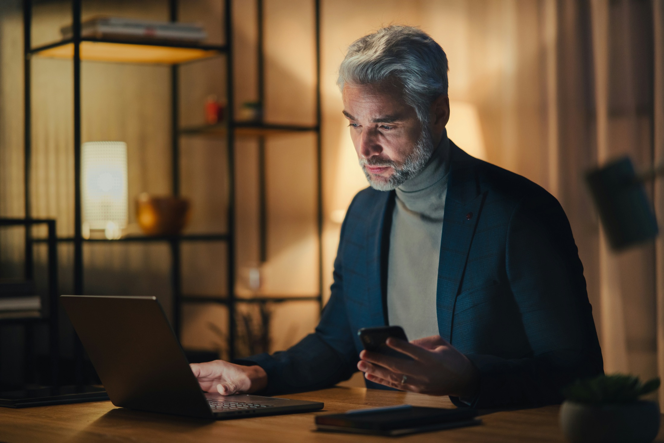 A man with gray hair works on a laptop while holding a smartphone, seemingly checking essential dates in a dimly lit room with shelves in the background.