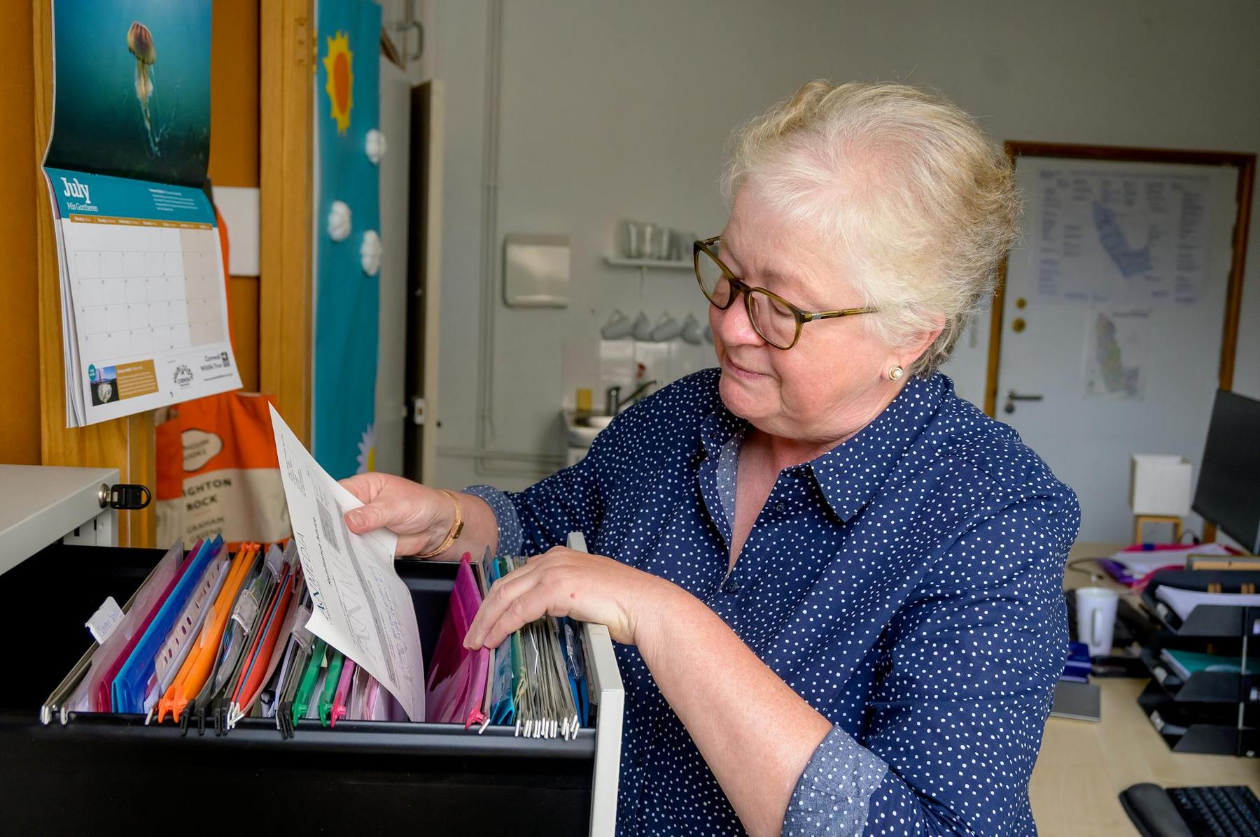 An older woman wearing glasses sorts through files in a filing cabinet in an office setting.