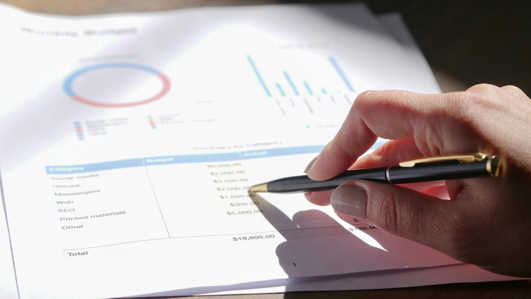A hand holding a pen points to figures on a printed business report with charts and a budget summary visible.