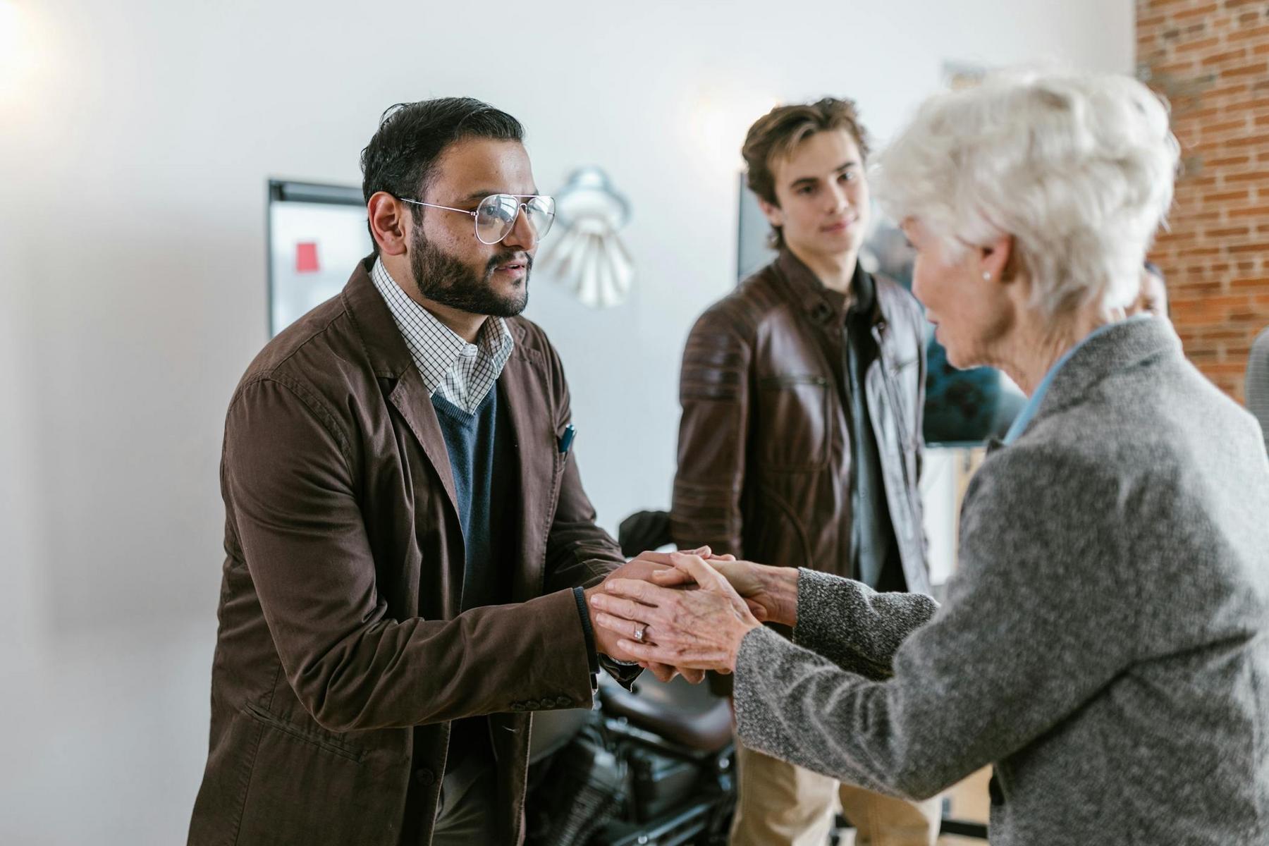A man in a brown jacket shakes hands with an older woman in a grey coat, while another man in a leather jacket stands nearby.