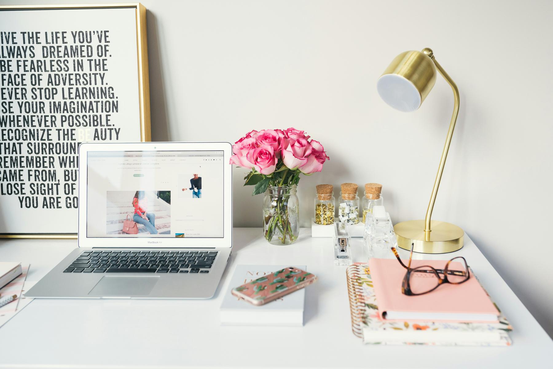 A tidy desk with a laptop, a vase of pink roses, a gold desk lamp, notebooks, glasses, a smartphone, and framed motivational quote against a light wall.