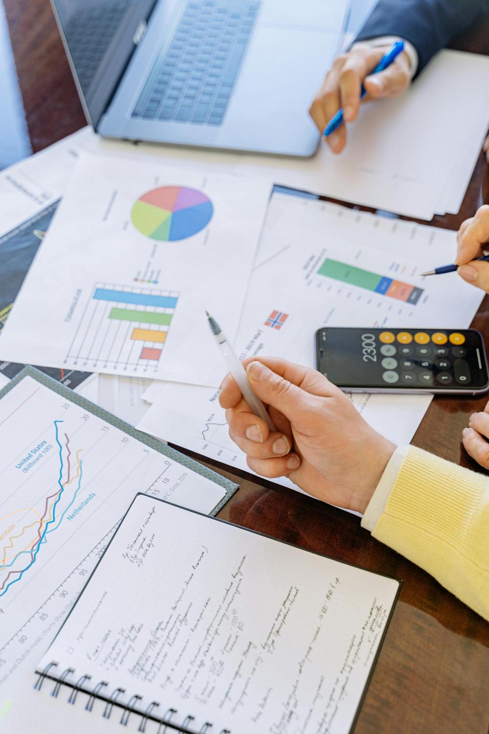 Two people work at a desk with charts, graphs, financial documents, a calculator, a notebook with notes, and a laptop.