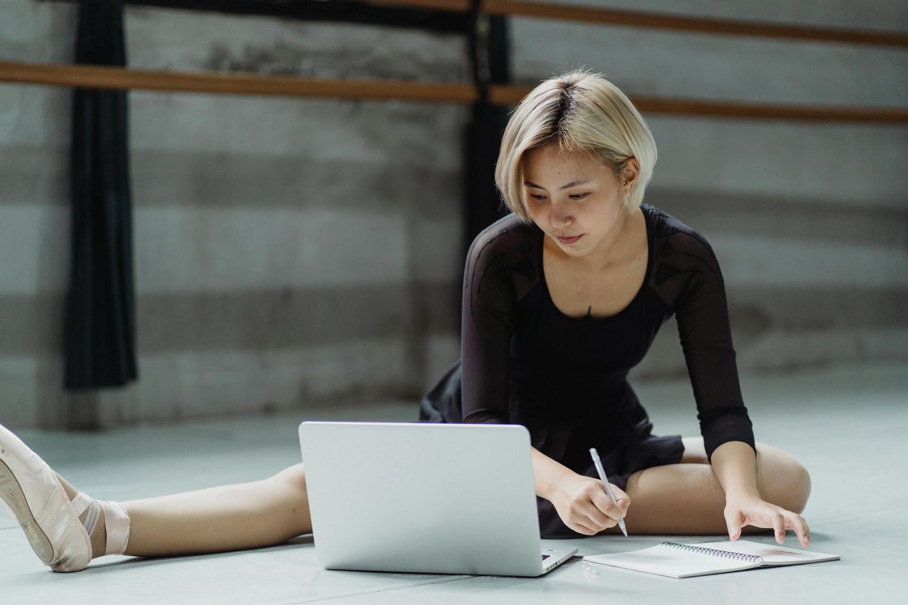 A person in ballet attire sits on the floor stretching, using a laptop and writing in a notebook inside a dance studio.