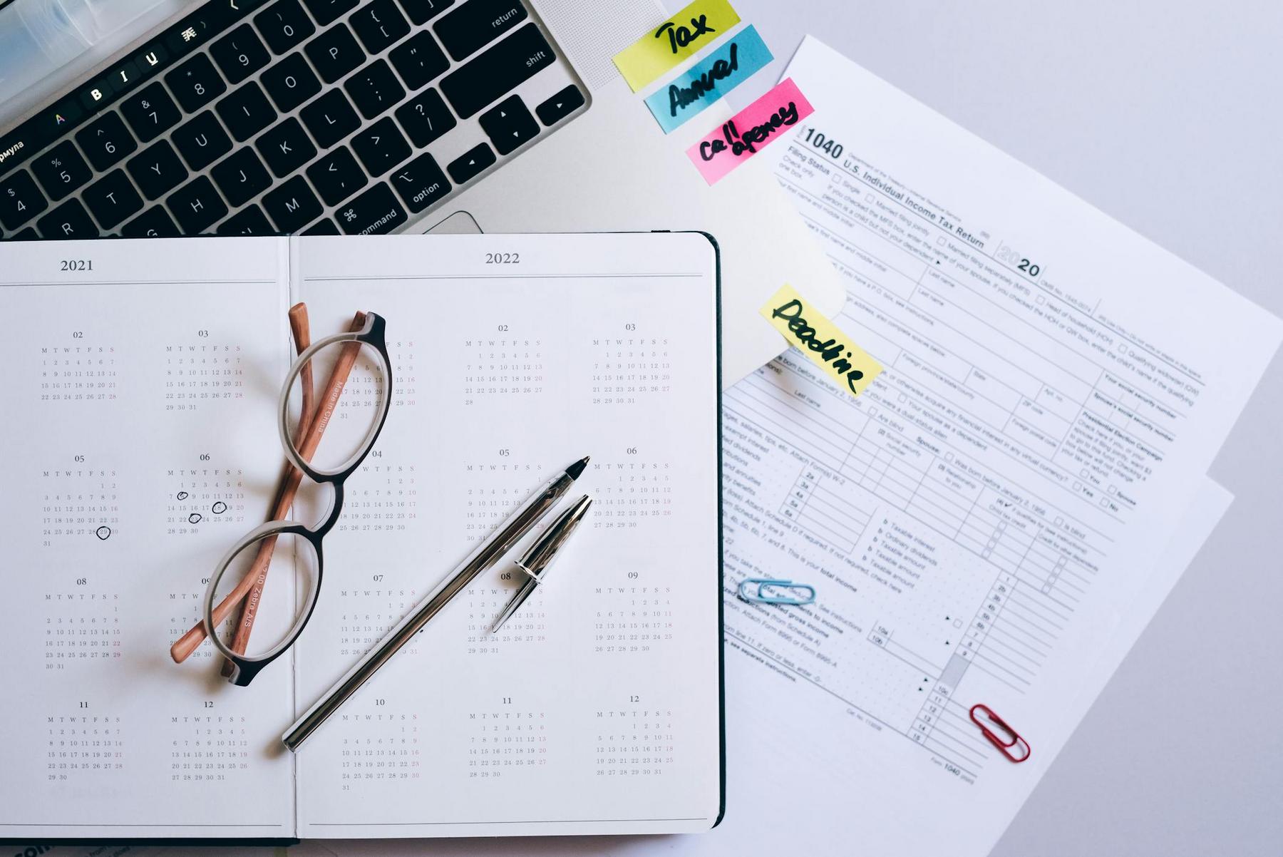 A planner, eyeglasses, and pens rest on a laptop keyboard next to tax forms and sticky notes labeled ‘Tax,’ ‘Annual,’ ‘Co. Report,’ and ‘Deadline.’.