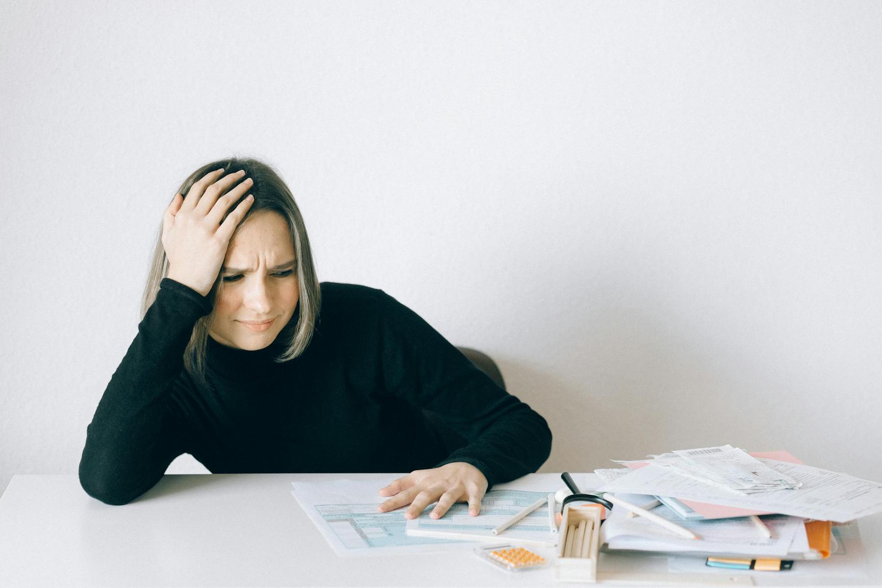 A woman sits at a table with her hand on her head, looking frustrated while reviewing documents and papers scattered in front of her.