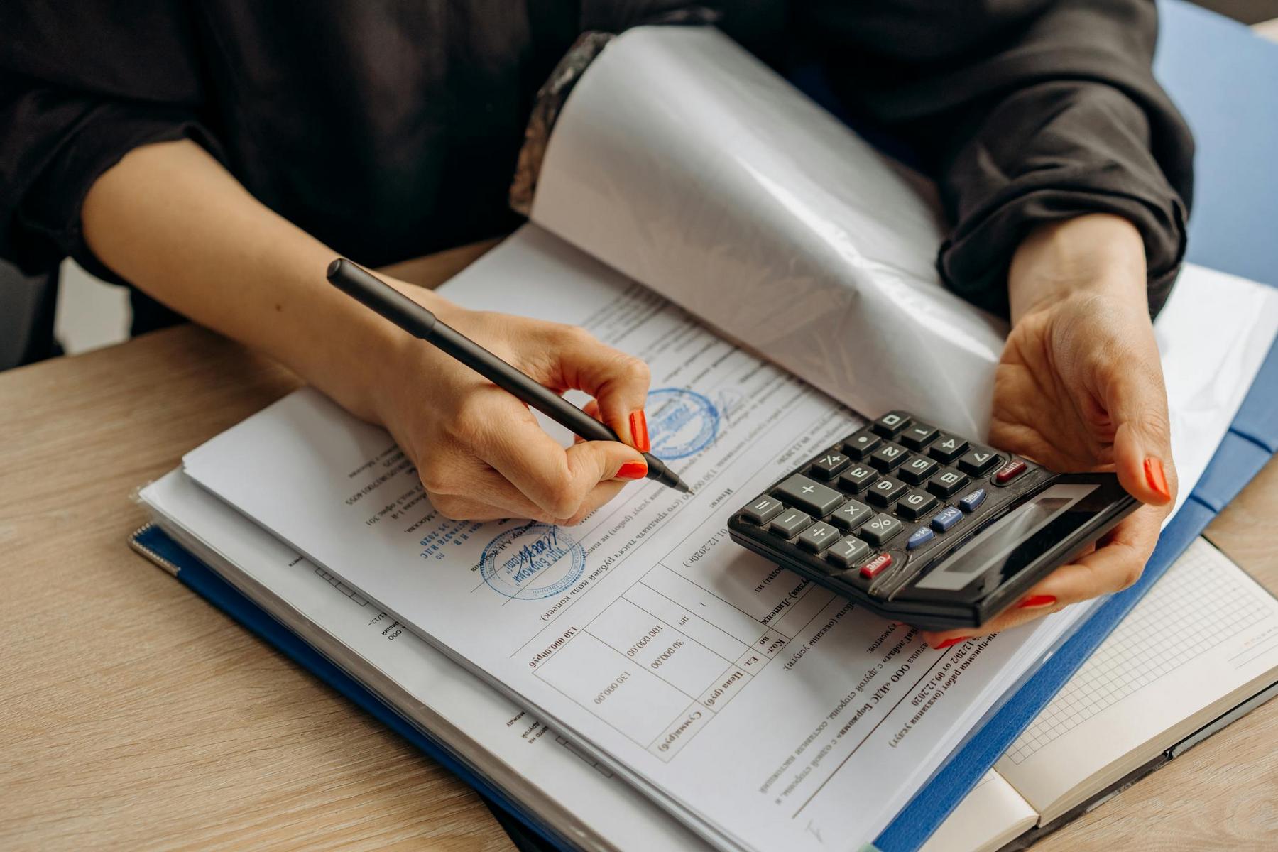 Person with red nail polish holding a calculator and writing on documents with charts and stamps at a desk.