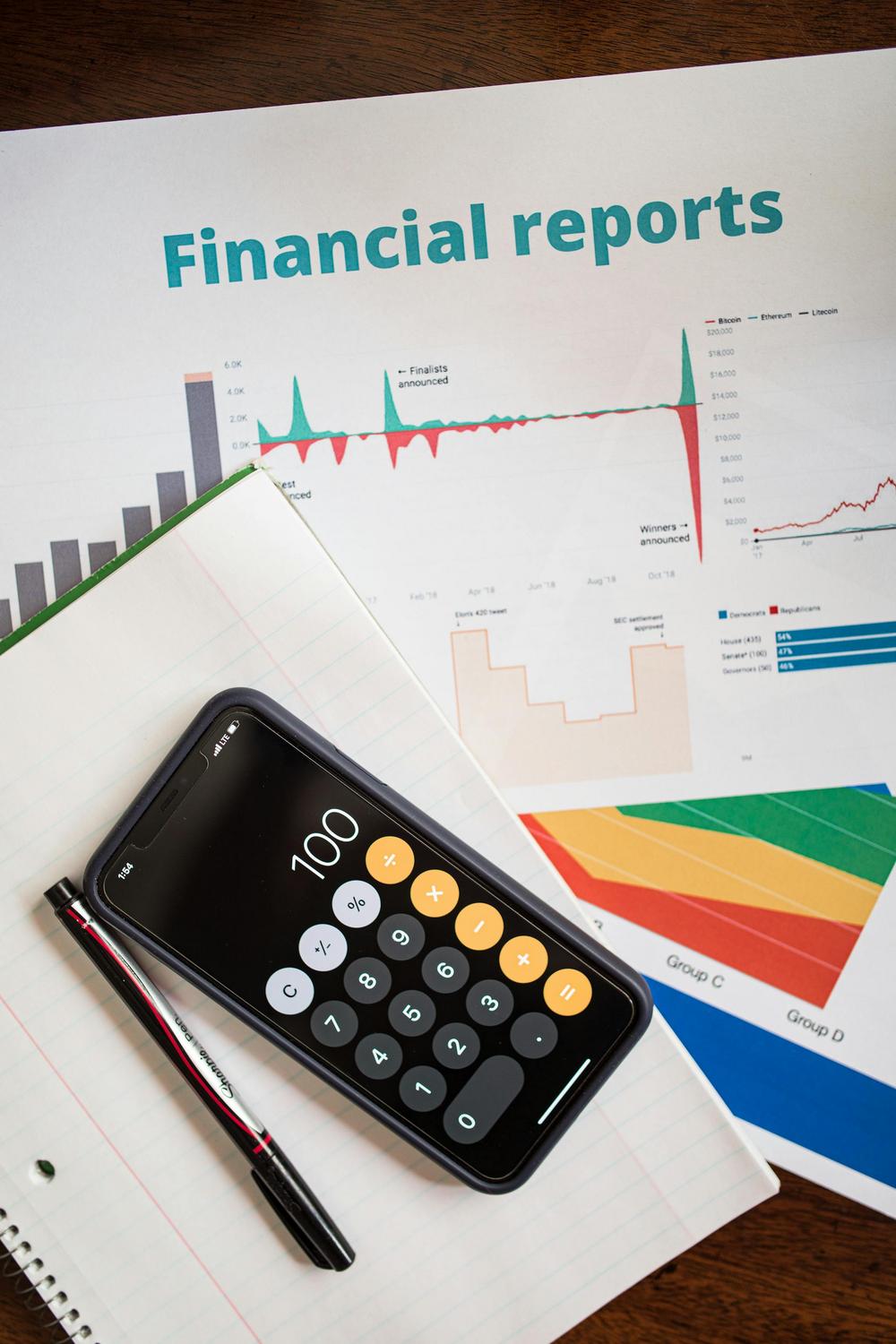 A smartphone with a calculator app, a pen, notebook, and printed financial reports with charts and graphs on a desk.