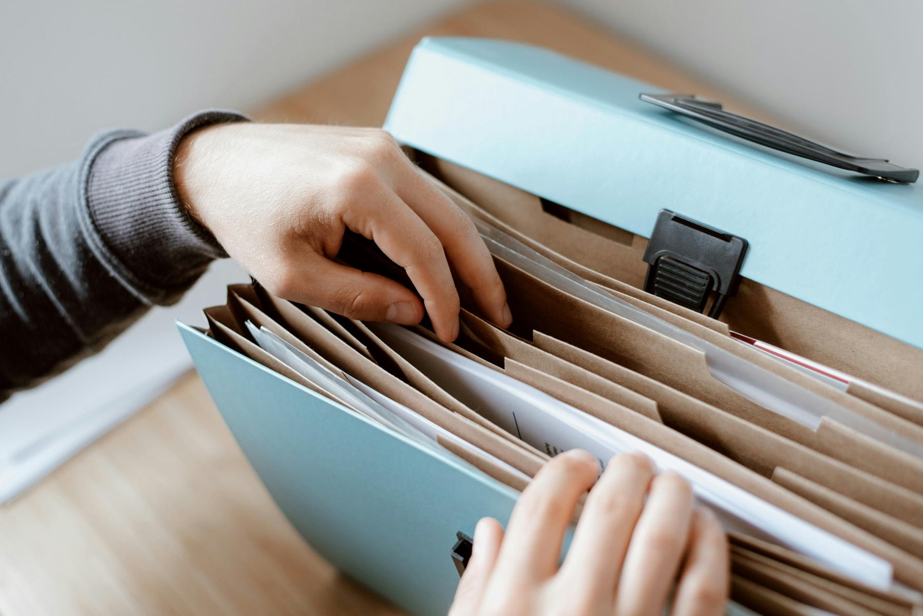 A person searches through documents organized in a blue accordion file folder on a wooden surface.