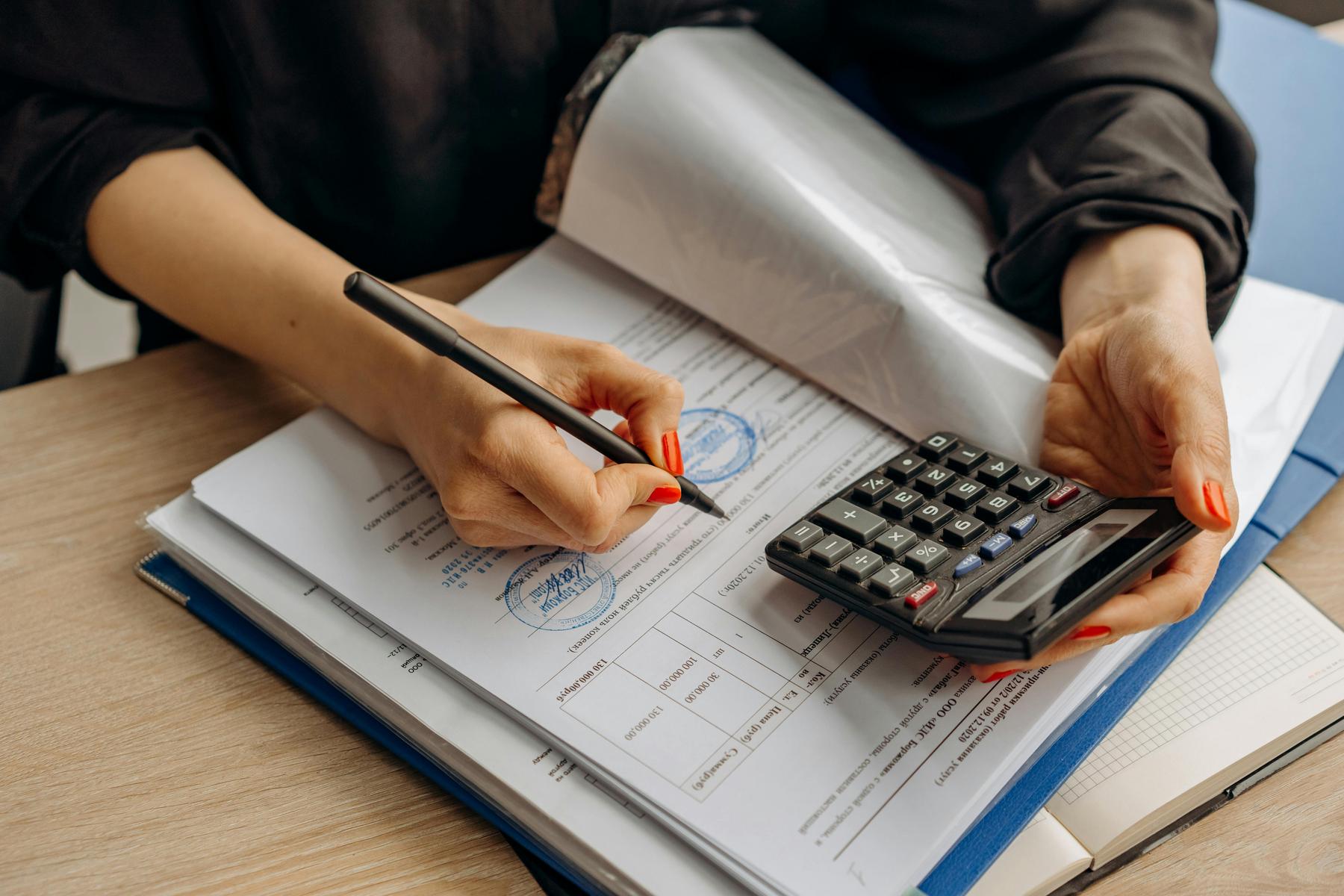 Person holding a calculator and pen, filling out documents on a desk with multiple folders and papers.