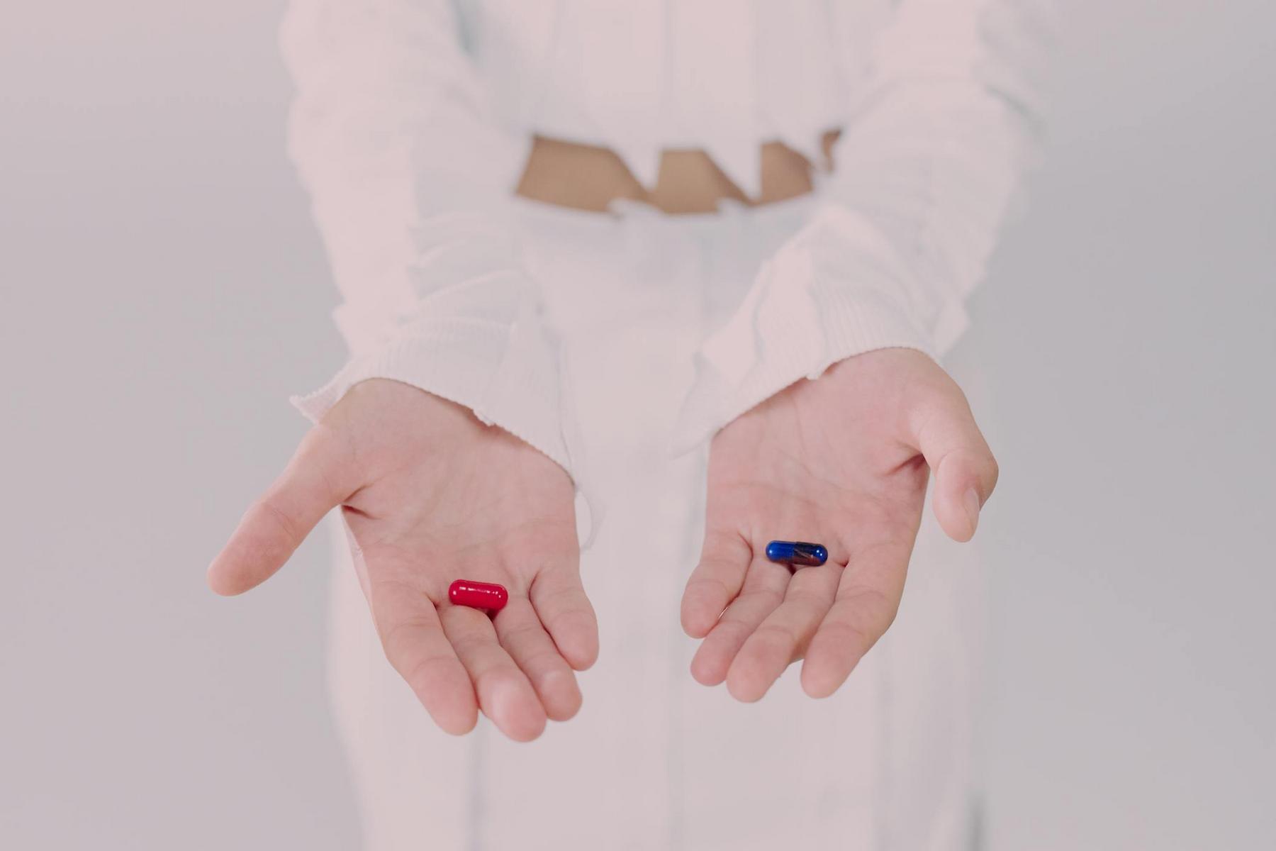 A person in white clothing holds a red capsule in their left hand and a blue capsule in their right hand, palms facing up.