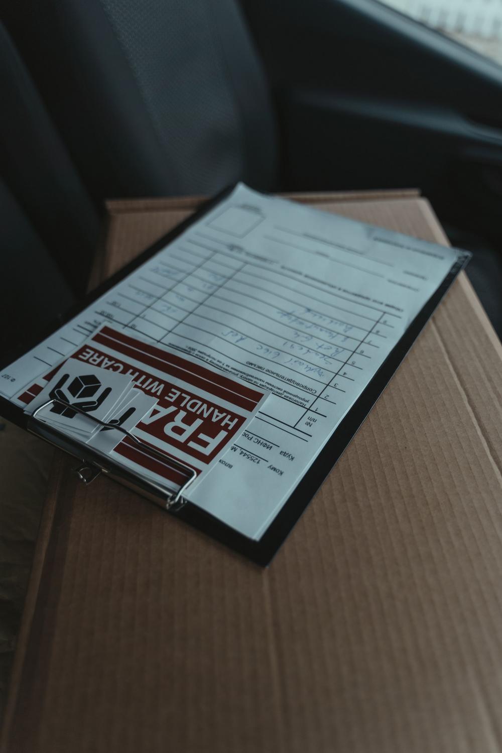 A clipboard with shipping documents and a "FRAGILE HANDLE WITH CARE" label rests on top of a cardboard box on a car seat.
