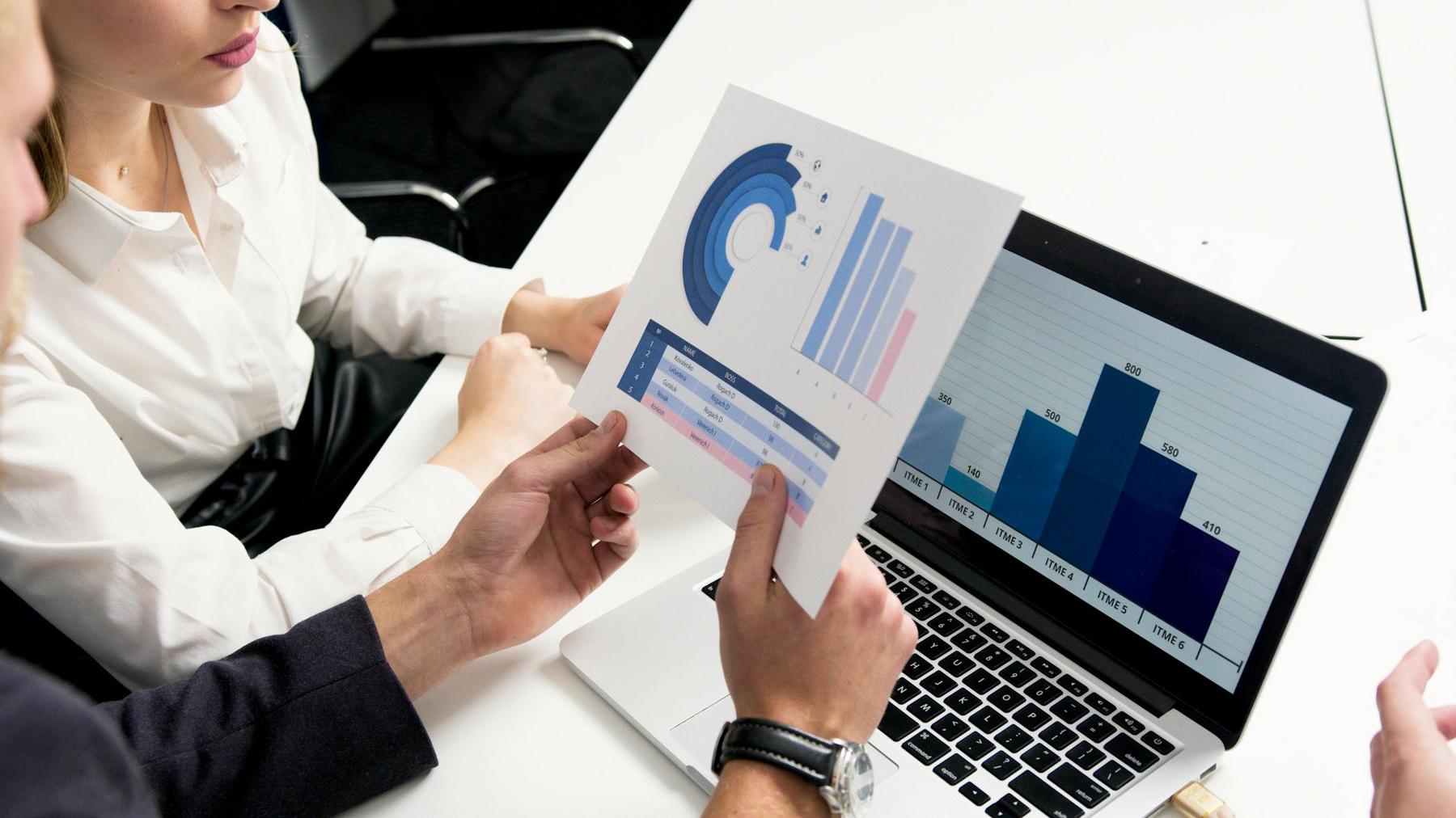 Two people review printed charts and a bar graph displayed on a laptop screen during a business meeting at a white table.