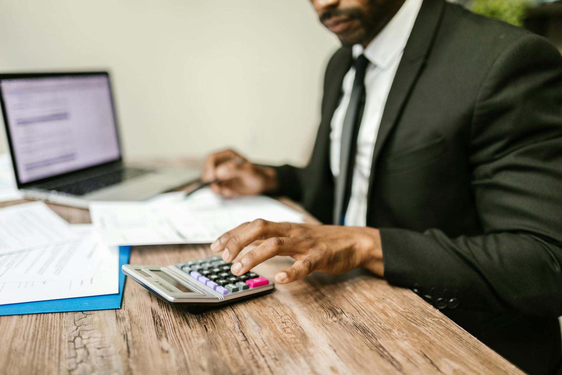A man in a suit uses a calculator at a desk with paperwork and a laptop.