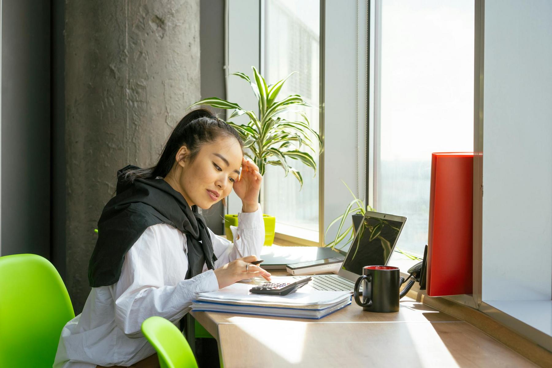 A woman sits at a desk by a window, using a calculator and looking at her laptop, with notebooks, a mug, and a plant nearby.