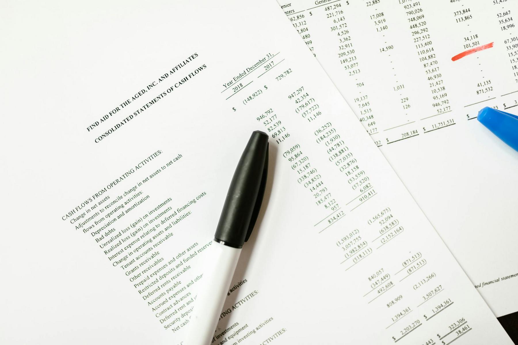 A black and white pen rests on financial statement documents with numbers and tables, next to a blue pen on a white surface.