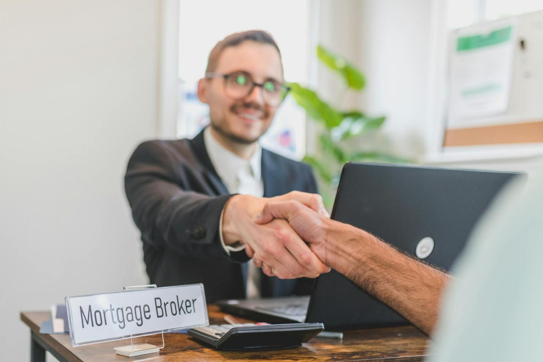 A person labeled as a mortgage broker sits at a desk, smiling and shaking hands with another individual across from them.
