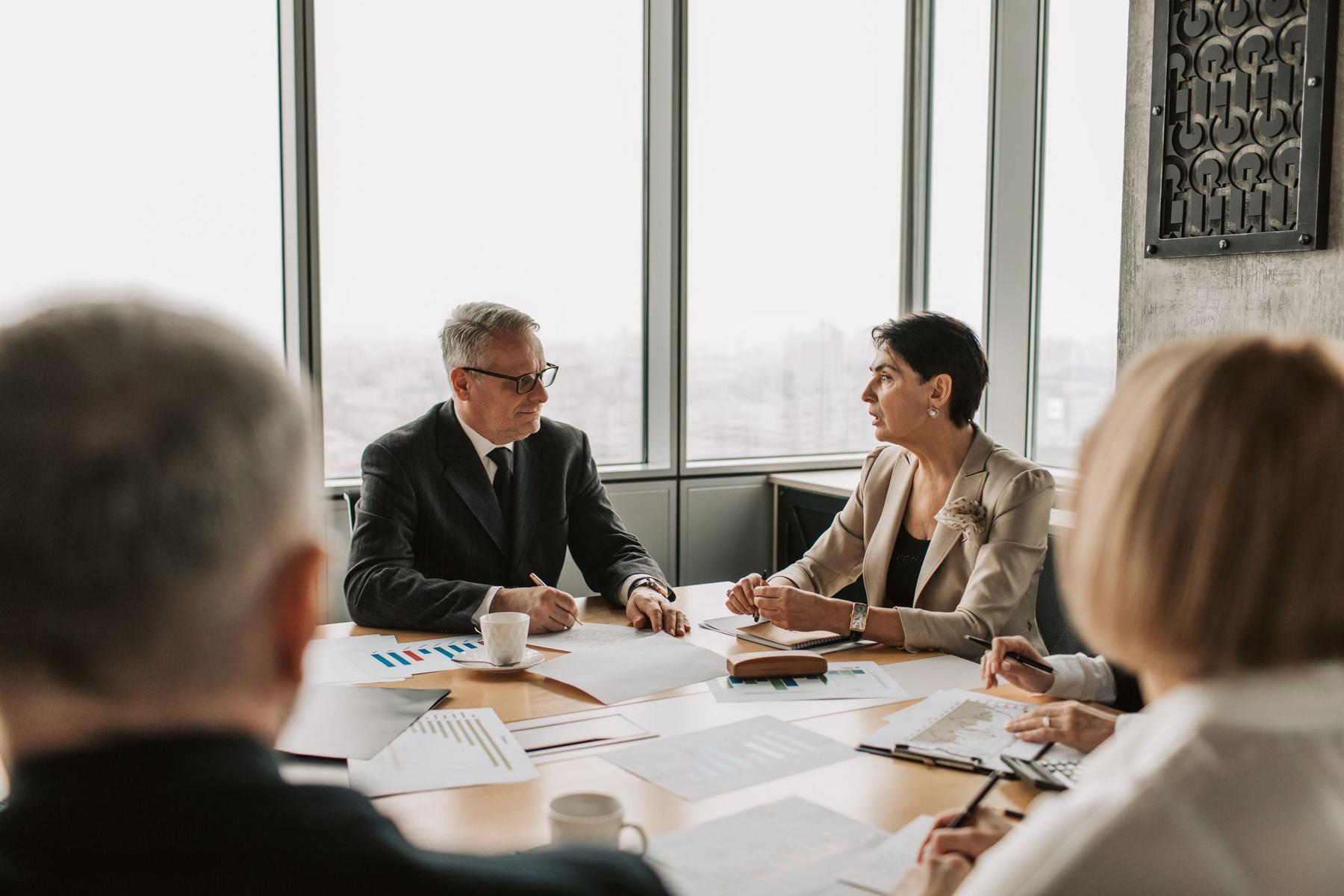 Four people in business attire sit around a conference table with documents, charts, and coffee cups, engaged in a meeting in a modern office with large windows.