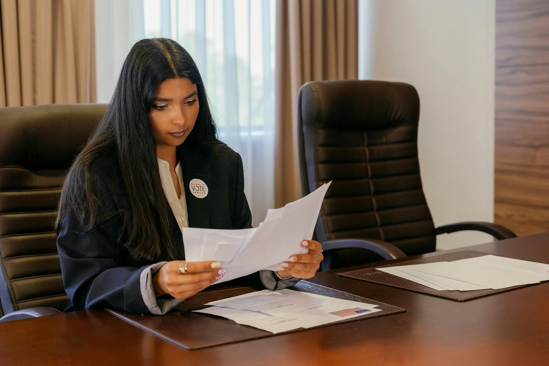 A woman in business attire sits at a conference table, reviewing documents. She wears a round button on her blazer. Two empty chairs are beside her.