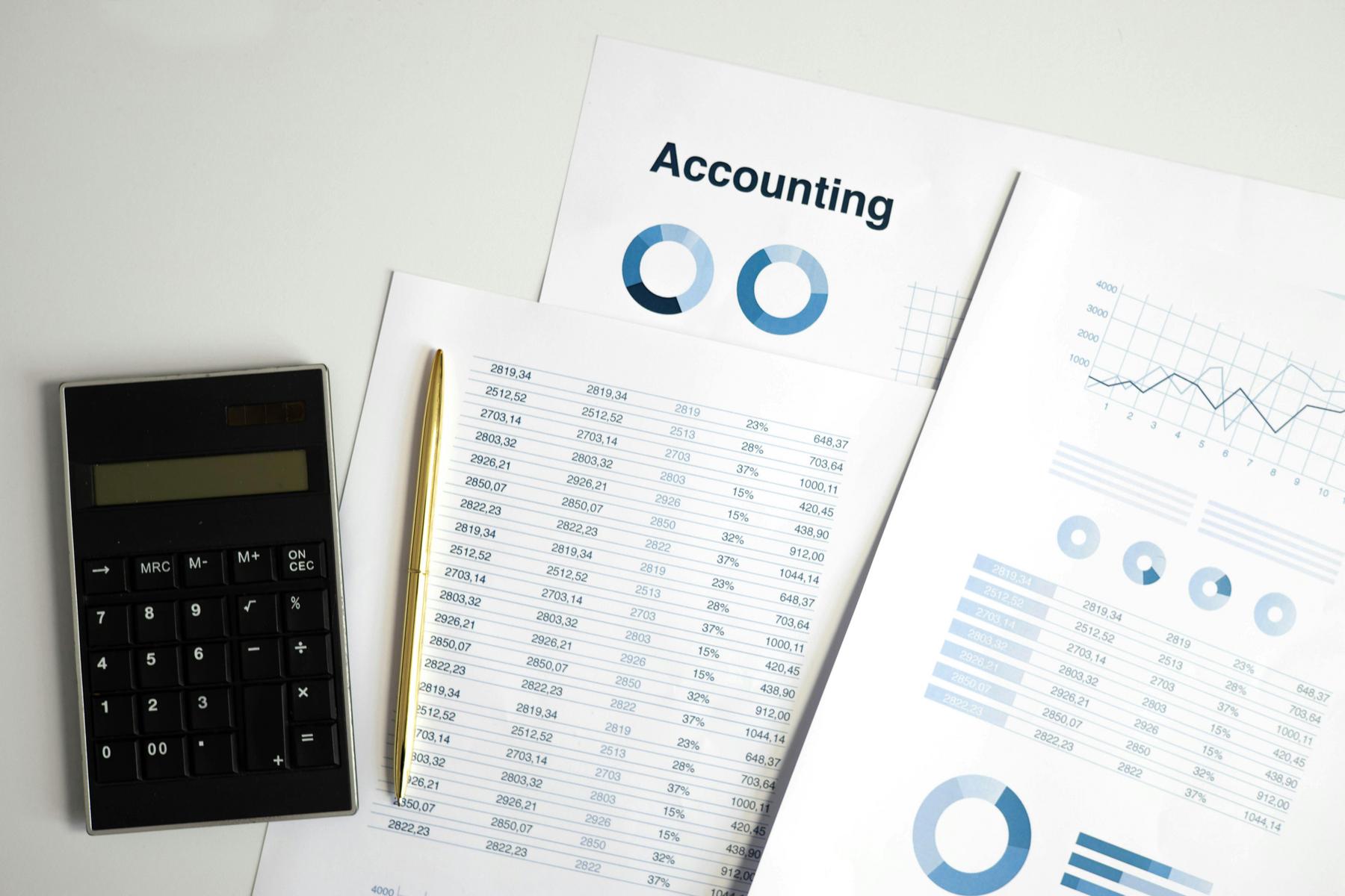 Calculator, pen, and printed accounting documents with charts, graphs, and data tables displayed on a white surface.